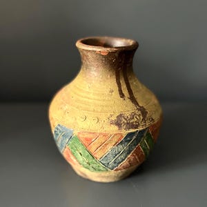 Puede incluir: Jarrón amarillo pequeño, hecho a mano, con cuerpo redondeado y cuello estrecho. El jarrón presenta un patrón geométrico en azul, verde y rojo. El borde y la parte superior del jarrón son de color marrón oscuro.