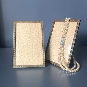 Puede incluir: Dos marcos de fotos rectangulares con bordes de metal en tono bronce que presentan un patrón geométrico repetitivo. Los marcos contienen inserciones con un patrón de piel sintética texturizada de color crema. Un collar de perlas de dos hilos descansa sobre uno de los marcos.