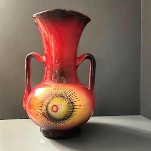 Op de afbeelding: Een keramische vaas met een levendig rood en oranje kleurverloop en twee zijhandvatten. Het lichaam toont een centraal cirkelvormig motief met een geel centrum, een rode stip en uitstralende zwarte lijnen op een donkere basis en rand.