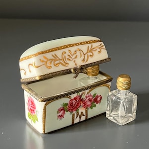 Pode incluir: Uma pequena caixa de porcelana aberta com tampa articulada, com um design floral de rosas e detalhes dourados. Um pequeno frasco de perfume de vidro transparente com tampa dourada está ao lado da caixa. A caixa e o frasco estão sobre uma superfície cinza.