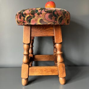 Op de afbeelding: Een houten kruk met vier gedraaide poten en een ronde zitting bekleed met een textielstof met een herhalend patroon van gebogen vormen in roze, gele, groene en zwarte tinten. Een rode en gele appel ligt bovenop de zitting van de kruk.