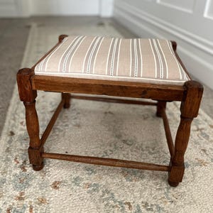 May include: A square footstool with a dark brown wooden frame featuring turned legs and a padded seat upholstered in a beige fabric with vertical white and dark brown stripes. The stool has a low profile and a simple, sturdy construction.