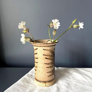 Może przedstawiać: Cylindryczny ceramiczny wazon o nakrapianym beżowym wykończeniu z ciemnobrązowymi wzorami przypominającymi liście. Wazon ma rozszerzany brzeg i zawiera kilka białych kwiatów z zielonymi łodygami oraz pękatymi kielichami.