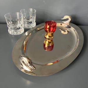 Könnte beinhalten: Ein ovales Serviertablett aus silberfarbenem Metall mit zwei schwanenförmigen Griffen, begleitet von zwei klaren Gläsern mit strukturiertem Boden und einem kleinen roten Schnapsglas mit goldfarbenem Boden auf der Tablettfläche.