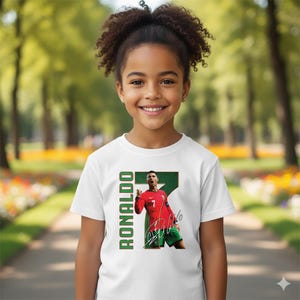 Op de afbeelding: Wit T-shirt met een afbeelding van een voetballer in een rood shirt, groene shorts en een handtekening. Het woord "RONALDO" staat verticaal in groen gedrukt. De speler viert een doelpunt.