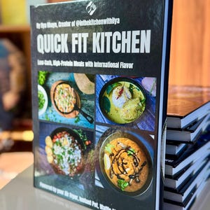 May include: A stack of cookbooks with a black cover. The top book's cover displays the title "QUICK FIT KITCHEN" in white. The cover also features images of various dishes and text that reads "Low-Carb, High-Protein Meals with International Flavour."
