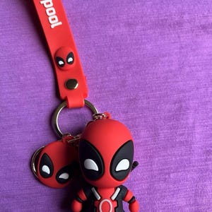 Puede incluir: Un llavero rojo de Deadpool con una pequeña figura de Deadpool y un dije con la cara de Deadpool. El llavero tiene una correa roja con la palabra "deadpool" en texto blanco.