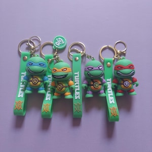 Könnte beinhalten: Vier grüne Schildkröten-Schlüsselanhänger mit goldfarbenen Metallringen und grünen Silikonbändern. Jedes Band zeigt den weißen Schriftzug TURTLES und ein orangefarbenes Schwert-Symbol. Die Figuren tragen Masken und Brustpanzer mit L, M, D und R.