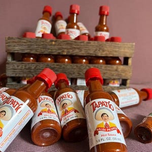 Op de afbeelding: Flessen Tapatio-hete saus met rode doppen en witte etiketten met een man in een sombrero. De flessen zijn gerangschikt in een houten krat en verspreid op een rood-wit gestreept oppervlak. Elke fles bevat 50 ml hete saus.