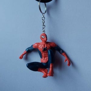 May include: A Spider-Man keychain featuring the superhero in a crouching pose. The figure is primarily red and blue, with a black spider emblem on the chest. The keychain includes a metal chain and a black ring.