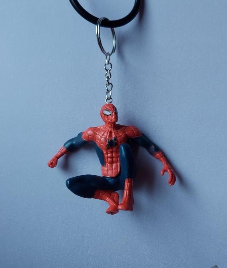 May include: A Spider-Man keychain featuring the superhero in a crouching pose. The figure is primarily red and blue, with a black spider emblem on the chest. The keychain includes a metal chain and a black ring.