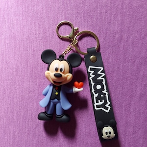 Op de afbeelding: Een Mickey Mouse sleutelhanger met een 3D-beeldje van Mickey in een blauw pak met een rood hart. De zwarte band leest "MICKEY" in het wit, met een Mickey Mouse-hoofd aan de onderkant. De sleutelhanger heeft een gouden ring.