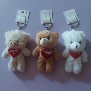 May include: Three plush teddy bear keychains in beige, light brown, and white. Each bear features a fuzzy texture, a small brown nose, and a red glittery heart-shaped patch on its chest with the text I LOVE YOU printed in white letters.