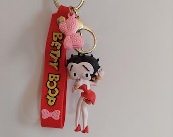Puede incluir: Un llavero de Betty Boop con una correa roja con la inscripción "BETTY BOOP" en letras amarillas. El llavero incluye una figura de Betty Boop con un vestido rojo y un charm de lazo rosa. Está unido a un anillo dorado.