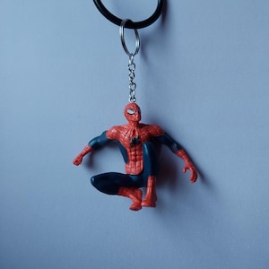 May include: A Spider-Man keychain featuring the superhero in a crouching pose. The figure is red and blue, with a black keychain and a silver ring. The background is a solid light blue.