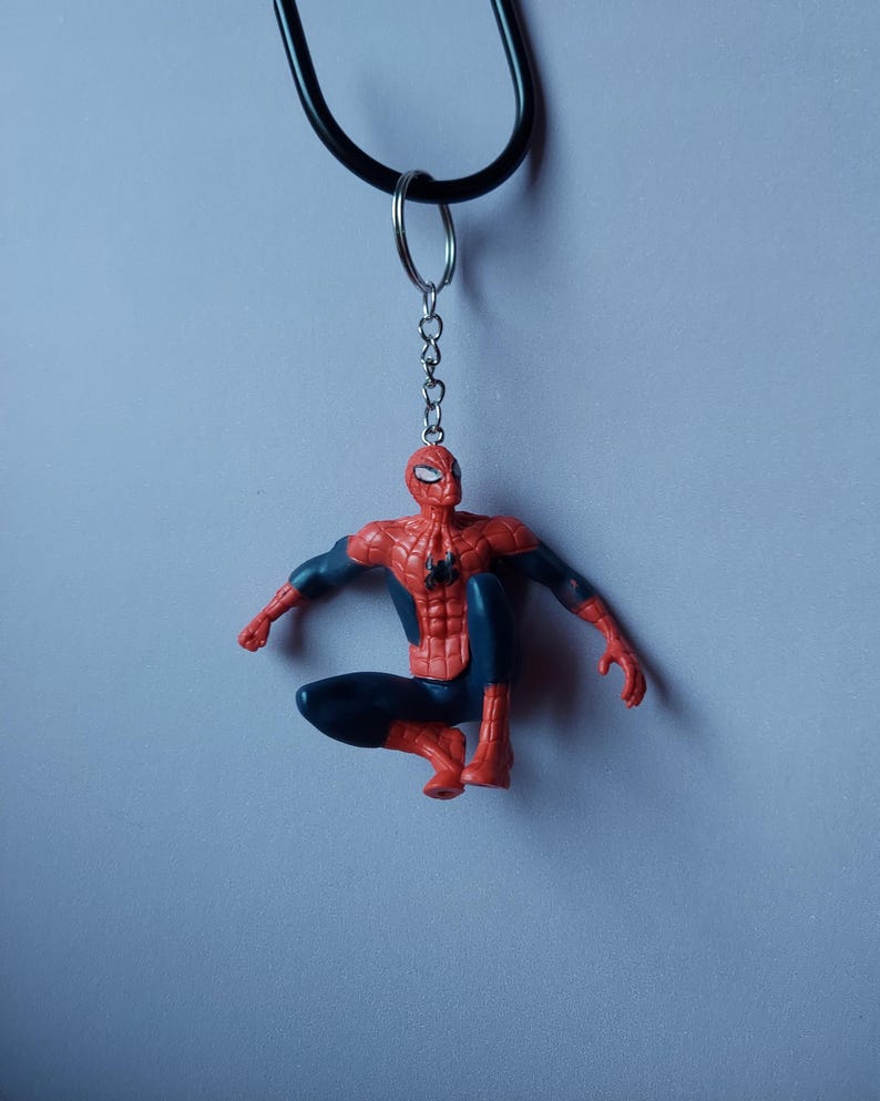 May include: A Spider-Man keychain featuring the superhero in a crouching pose. The figure is red and blue, with a black keychain and a silver ring. The background is a solid light blue.