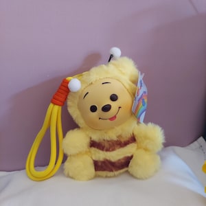Op de afbeelding: Een pluche Winnie de Poeh verkleed als een bij. Het speelgoed is geel met bruine strepen en een lachend gezicht. Het heeft een gele lus met een oranje accent en een label met een regenboog.
