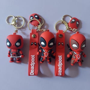 Puede incluir: Tres llaveros de Deadpool con figuras rojas y negras de ojos blancos. Dos llaveros incluyen una correa de silicona roja con el texto Deadpool impreso en blanco, y los tres cuentan con anillas y clips de metal en tono dorado.