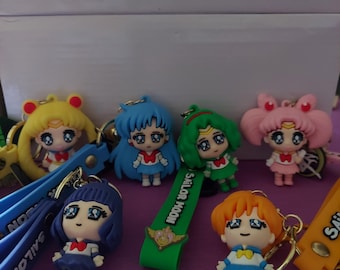 Op de afbeelding: Een verzameling kleurrijke Sailor Moon-personage sleutelhangers. Elke sleutelhanger heeft een miniatuurfiguur met verschillende haar- en outfitkleuren, waaronder geel, blauw, groen en roze. Sommige sleutelhangers hebben lanyards met de tekst "Sailor Moon".