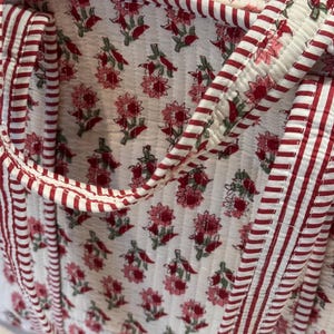 May include: Close-up of a quilted tote bag with a floral pattern. The bag features a white background with a repeating pattern of pink flowers and green leaves. The edges and handles are trimmed with red and white striped fabric.