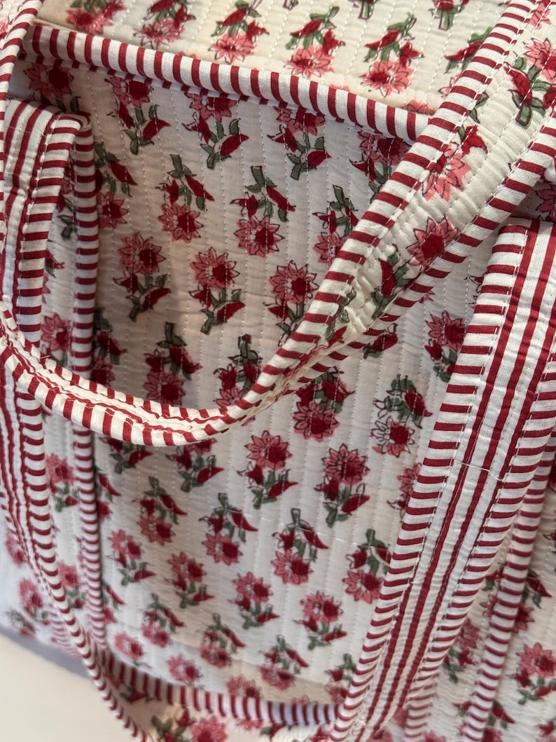 May include: Close-up of a quilted tote bag with a floral pattern. The bag features a white background with a repeating pattern of pink flowers and green leaves. The edges and handles are trimmed with red and white striped fabric.