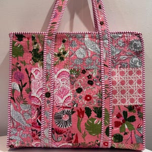 May include: Pink tote bag with a patchwork design featuring floral and bird patterns. The bag has a rectangular shape with a quilted texture and pink and white striped handles and trim. The bag is made of fabric.