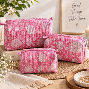 May include: A set of three rectangular quilted cosmetic bags in bright pink with a white floral pattern. Each bag features a zip closure and a structured shape. The bags vary in size, with one large, one medium, and one small pouch.