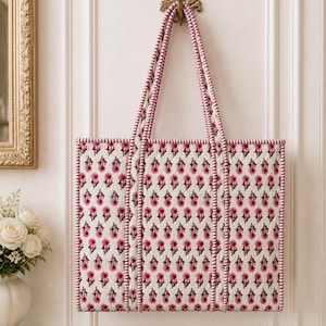 May include: A rectangular tote bag with a floral pattern in pink and white hangs from a decorative gold hook. The bag has pink and white striped handles and trim. The floral pattern is repeated across the bag's surface.