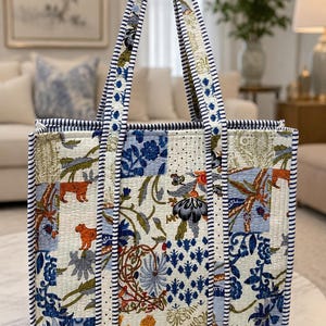 May include: A quilted tote bag with a patchwork design featuring floral and animal motifs in blue, orange, and cream. The bag has sturdy handles and is trimmed with a navy and white striped border.