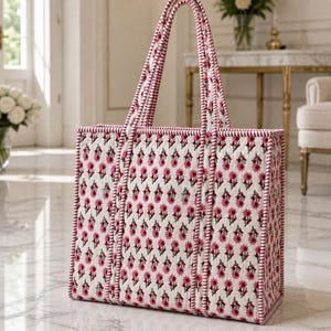 May include: A rectangular tote bag with a white background and a repeating pattern of small pink flowers. The bag has pink handles and trim. The bag is made of a quilted fabric.
