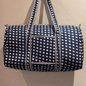 May include: A navy and white checked duffel bag with white trim and handles. The bag is rectangular with a front pocket and a zip closure. The handles are padded for comfort.