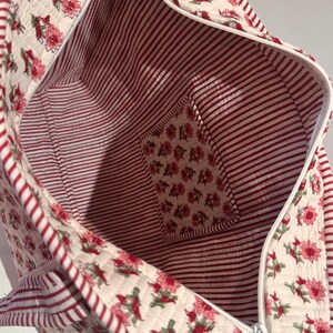 May include: Open view of a quilted tote bag. The exterior features a cream background with a floral pattern in shades of red. The interior has red and white stripes, with a small pocket featuring the floral pattern. A white zipper runs along the top.
