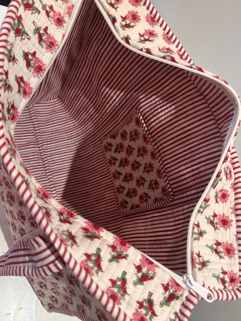 May include: Open view of a quilted tote bag. The exterior features a cream background with a floral pattern in shades of red. The interior has red and white stripes, with a small pocket featuring the floral pattern. A white zipper runs along the top.