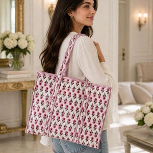 May include: A white tote bag with a pink floral pattern and pink trim. The bag has two handles and is being carried over the shoulder. The background is a well-lit room with a mirror and flowers.