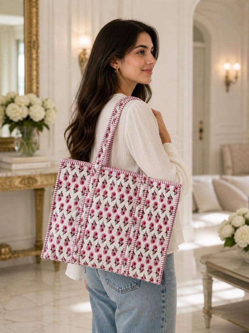 May include: A white tote bag with a pink floral pattern and pink trim. The bag has two handles and is being carried over the shoulder. The background is a well-lit room with a mirror and flowers.