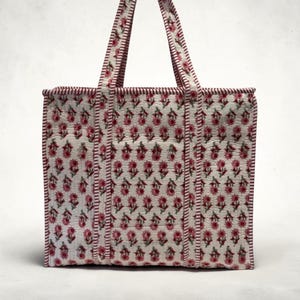 May include: A white tote bag with a repeating floral pattern in pink and green. The bag has two handles and is trimmed with a dark red stitching. The floral pattern is evenly distributed across the bag's surface.