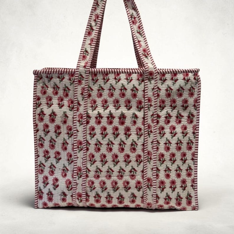 May include: A white tote bag with a repeating floral pattern in pink and green. The bag has two handles and is trimmed with a dark red stitching. The floral pattern is evenly distributed across the bag's surface.
