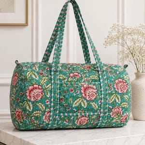 May include: A quilted duffel bag featuring a teal green base with a repeating floral pattern in shades of red, pink, and white. The bag has two long shoulder straps with a striped trim and a rectangular exterior pocket on the front side.