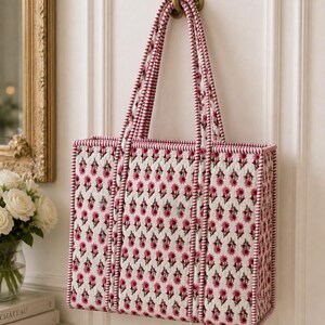 May include: A rectangular tote bag with a floral pattern in pink and white, hanging from a decorative brass hook. The bag has matching pink and white striped handles and trim. The floral pattern is repeated across the bag's surface.