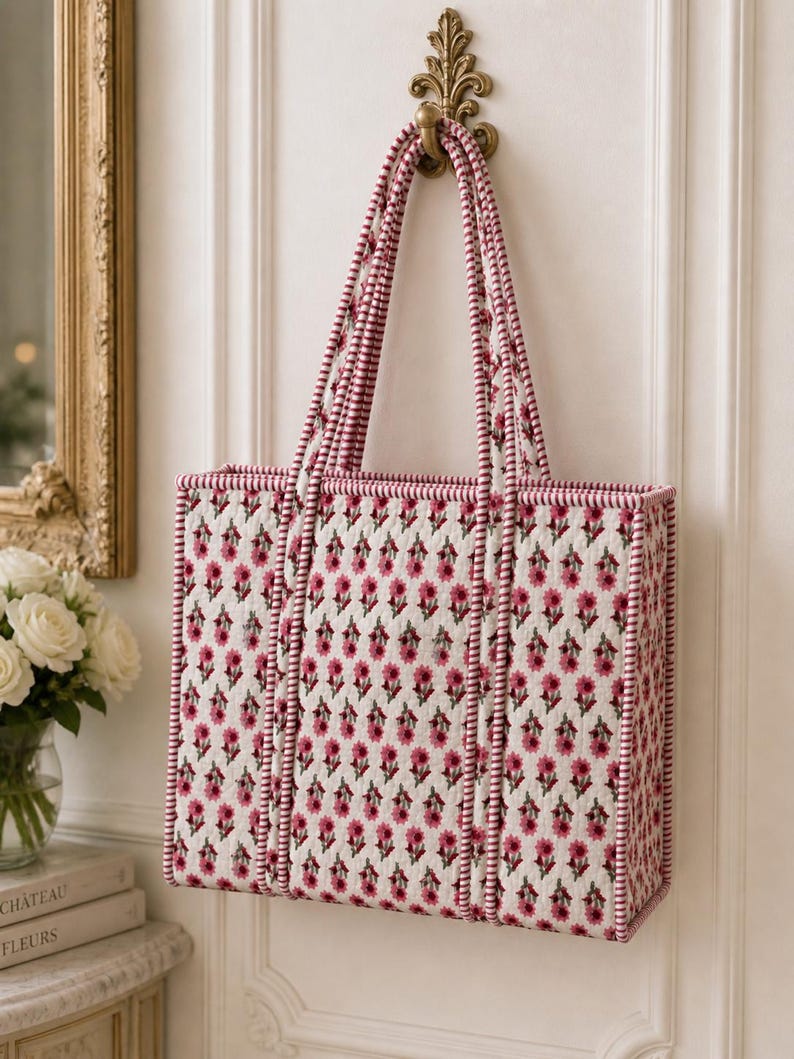 May include: A rectangular tote bag with a floral pattern in pink and white, hanging from a decorative brass hook. The bag has matching pink and white striped handles and trim. The floral pattern is repeated across the bag's surface.
