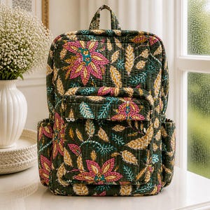 May include: A quilted backpack with a floral pattern in shades of pink, yellow, and teal on a dark green background. The backpack has a top handle, a front pocket, and side pockets. The bag is approximately 40 cm tall.