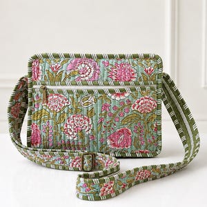 May include: A quilted crossbody bag featuring a light green background with a pink and dark green floral pattern. The bag has a rectangular shape, a zippered front pocket, and an adjustable shoulder strap with a green and white striped trim.