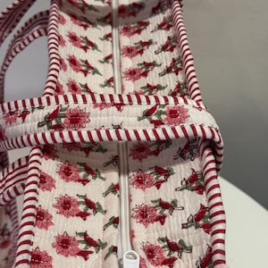 May include: A quilted tote bag with a floral pattern in pink and red on an off-white background. The bag has a white zipper and red and white striped trim and handles. The bag is designed for carrying items.