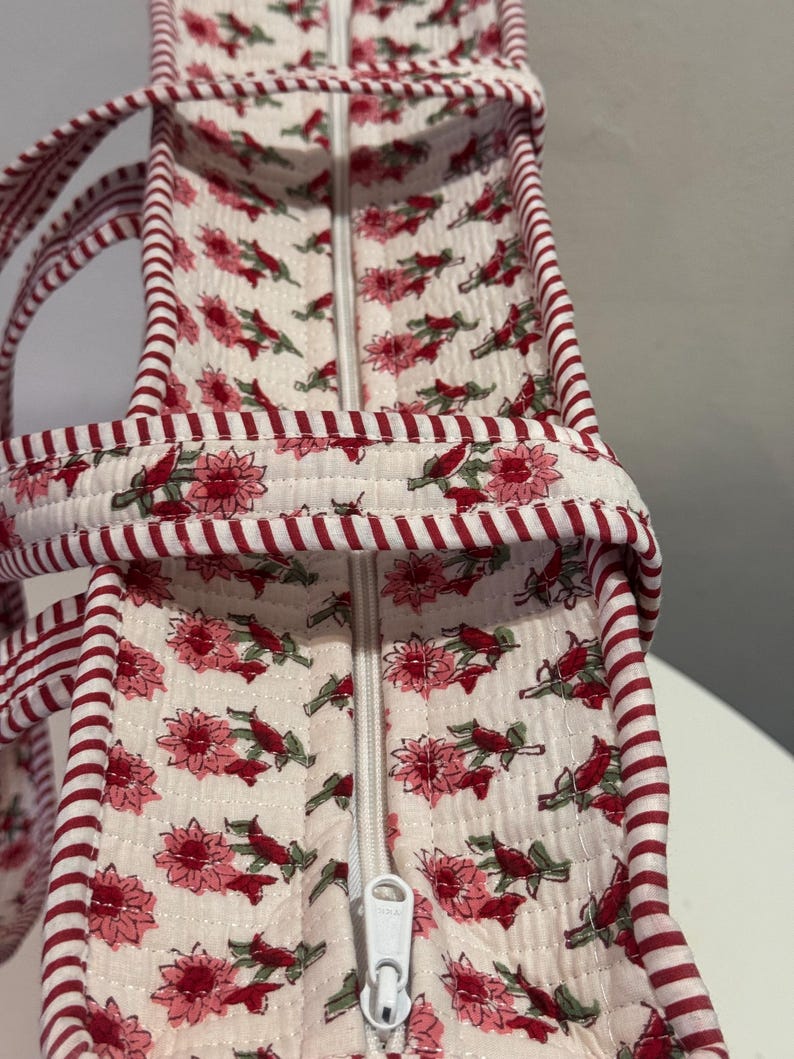 May include: A quilted tote bag with a floral pattern in pink and red on an off-white background. The bag has a white zipper and red and white striped trim and handles. The bag is designed for carrying items.