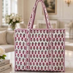 May include: A white tote bag with a floral pattern in shades of pink and green. The bag has a rectangular shape with matching pink handles and trim. The bag is sitting on a marble table.