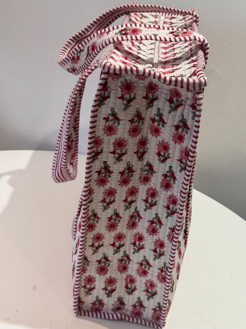May include: A quilted tote bag with a floral pattern in pink and red on a white background. The bag has a rectangular shape with a zipper closure and matching handles. The edges are trimmed with a red and white striped fabric.