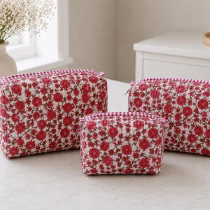 May include: A set of three rectangular quilted pouches in varying sizes, featuring a white background with a dense pattern of red flowers and green stems. Each pouch has a zip closure with a red and white chequered fabric trim along the top edge.