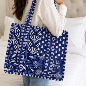 May include: A rectangular quilted tote bag featuring a patchwork design of deep blue and white patterns, including stylised leaves, raindrops, and concentric circles. The bag has long shoulder straps with a blue and white striped fabric trim.