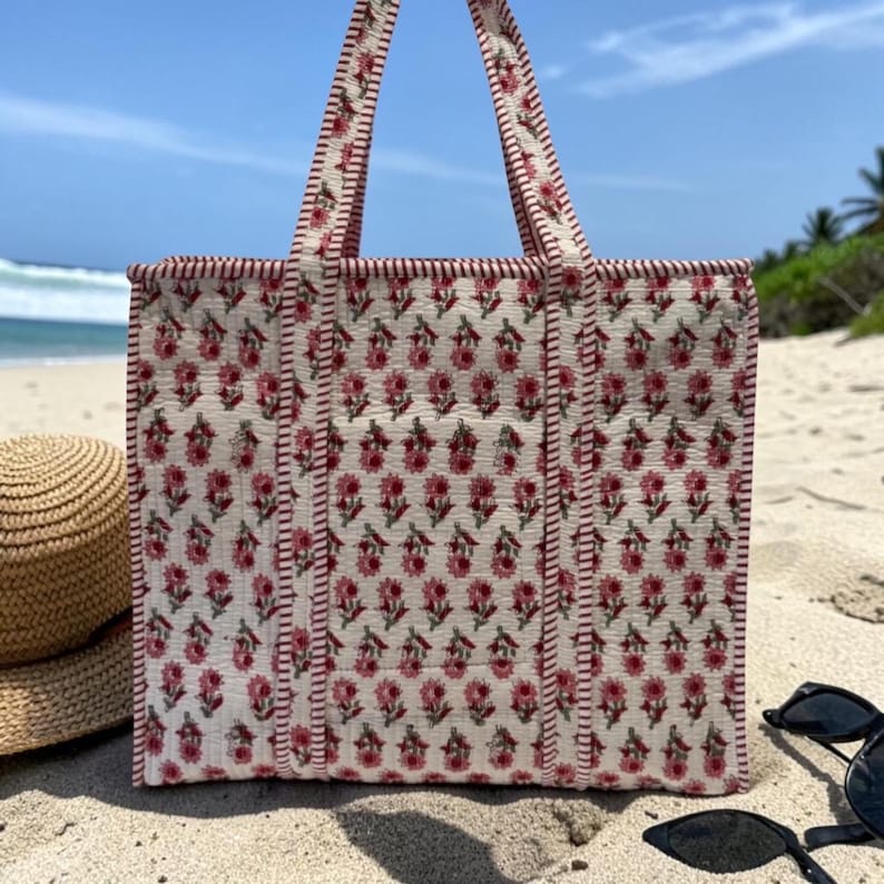 May include: A white tote bag with a repeating floral pattern in red and green. The bag has red trim and handles, and is sitting on a sandy beach. A straw hat and sunglasses are also visible.
