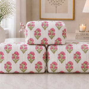 May include: Three rectangular quilted cosmetic bags featuring a white base with a repeating pink and green floral block print pattern. Each bag has a soft, ribbed texture and a small fabric tie attached to the side zipper pull for easy opening.